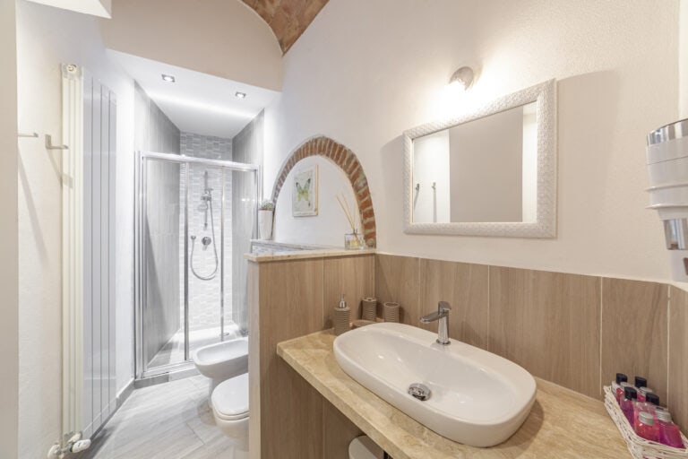 Bagno moderno con ampia doccia in vetro, lavabo in ceramica con specchio decorato, e dettagli in legno e mattoni a vista per un tocco di eleganza rustica.