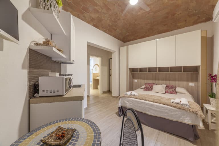 Monolocale elegante con soffitto in mattoni a volta, letto matrimoniale con armadi a ponte, angolo cottura attrezzato e tavolino con sedie. Ingresso al bagno con arredo moderno.