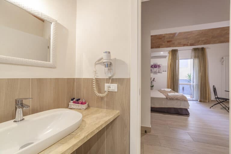 Bagno con lavabo moderno, specchio decorato e asciugacapelli a parete. Vista sulla camera con soffitto in mattoni a volta, letto matrimoniale e accesso al cortile.