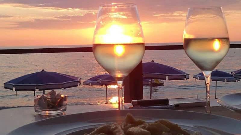 Cena al tramonto con due calici di vino bianco, un piatto di pasta e vista sul mare con ombrelloni blu.